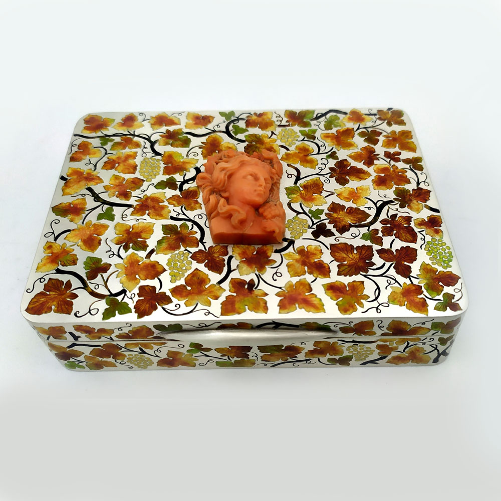 3813-4238 - Rectangular table box with rounded corners in 925/1000 sterling silver brossè with very fine hand engraving, also on the sides, of “ramage” with leaves and bunches of grapes fire-enamelled with various autumn colours, in Art Nouveau style. Beautiful handmade coral sculpture depicting a woman's face, in the center of the lid. Dimensions cm. 9 x 12 x 2.8. Weight gr. 330. Coral mm. 28 x 37. Lost hinge, i.e. semi-invisible. Designed by Giorgio Salimbeni in 1965 on inspiration from models previously made by the Salimbeni firm and manufactured in Florence in various specimens with different subjects and colours, again in the Salimbeni firm's headquarters with completely manual execution by artisan artists with thick slabs and large suitable reinforcements to sustain numerous enamelled firings at high heat at around 750° < 800° C.